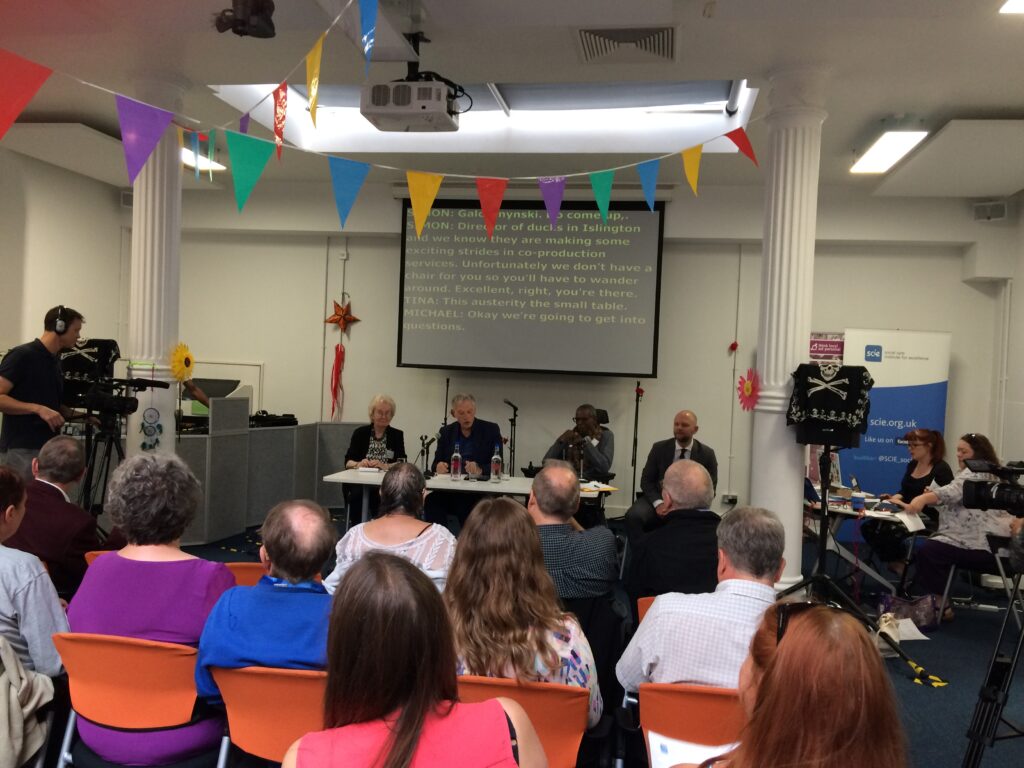 Photo of people at an event. The photo is taken from the back of the room and shows lines of people sitting on chairs, facing towards 4 people who are at the front of the meeting.