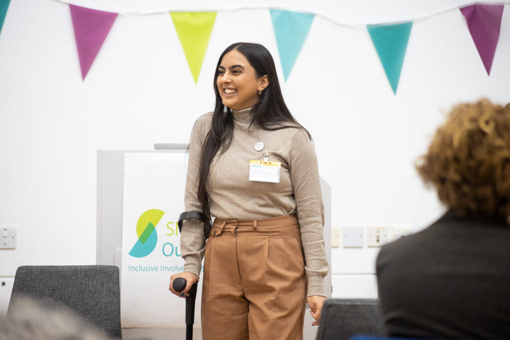 Image shows Suhkjeen Kaur from Chronically Brown. She is a young Asian woman using a crutch speaking at the Shaping Our Lives conference.