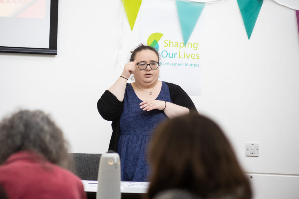 An image of Deidre from Zebra Access presenting at the conference. She is a woman standing in front of a poster with the Shaping Our Lives logo, and she is signing using British Sign Language.