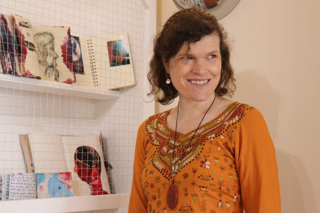 Image of Lorna Collins, a woman standing before some shelves of books. She is wearing an orange top and a long necklace, looking slightly to her left, with a smile.