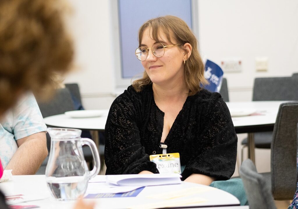 An image of Roxane Lavanchy - a young woman with brown hair and glasses. She is sitting at a table looking slightly to her right, smiling.