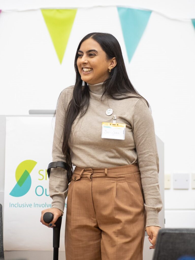 An image of Sukhjeen Kaur, founder of Chronically Brown. She is a young Asian woman standing in front of the Shaping Our Lives logo. She is using a walking aid, and is looking to her right, smiling.