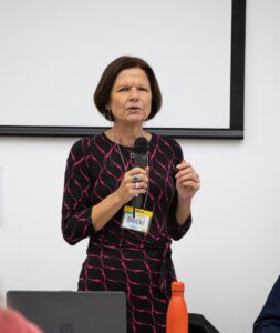 An image of Becki Meakin - a white woman with short brown hair. She is standing, holding a microphone in front of a laptop and a white board.