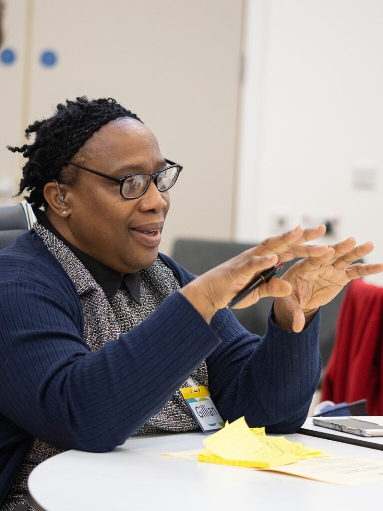An image of Gillian - a black woman with black-framed glasses and a hearing aid, who is sitting at a table, with yellow post it notes in front of her. She is holding a pen in her right hand and is gesturing with her hands while talking.
