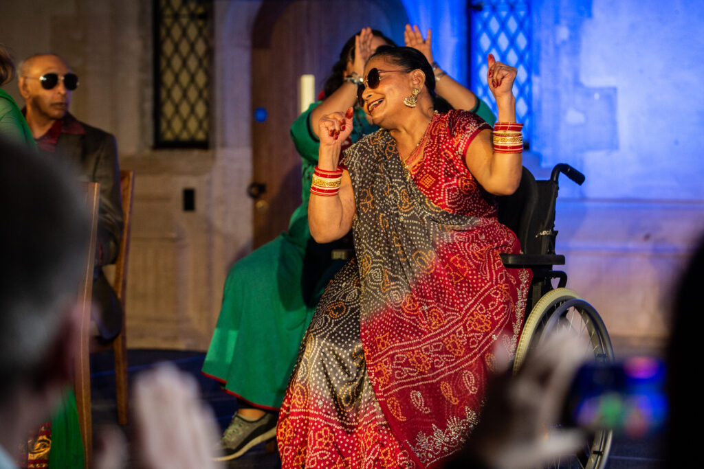 A photo of an older Asian woman. She is wearing a red patterned sari, multiple bangles, and sunglasses, and is holding her arms in the air, a smile on her face. She is a wheelchair user.