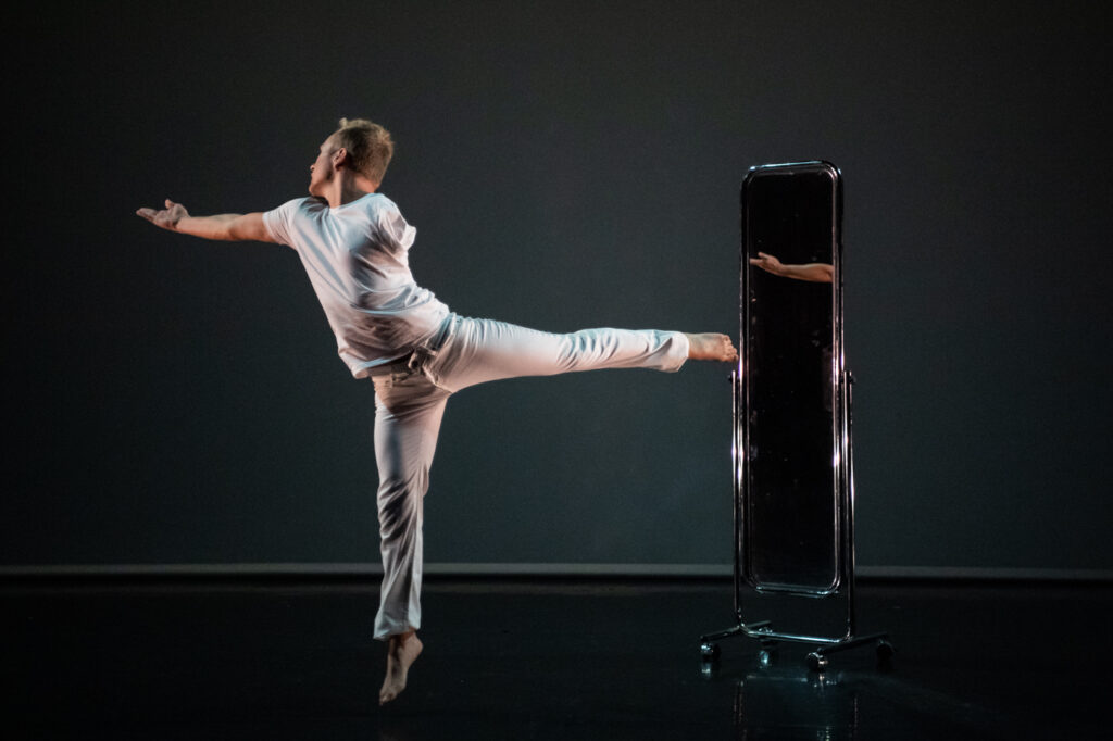 A photo of a man dancing. He is dressed all in white, and has one arm. His left leg is extended behind him, his right arm stretched before him, and behind his back there is a freestanding mirror.