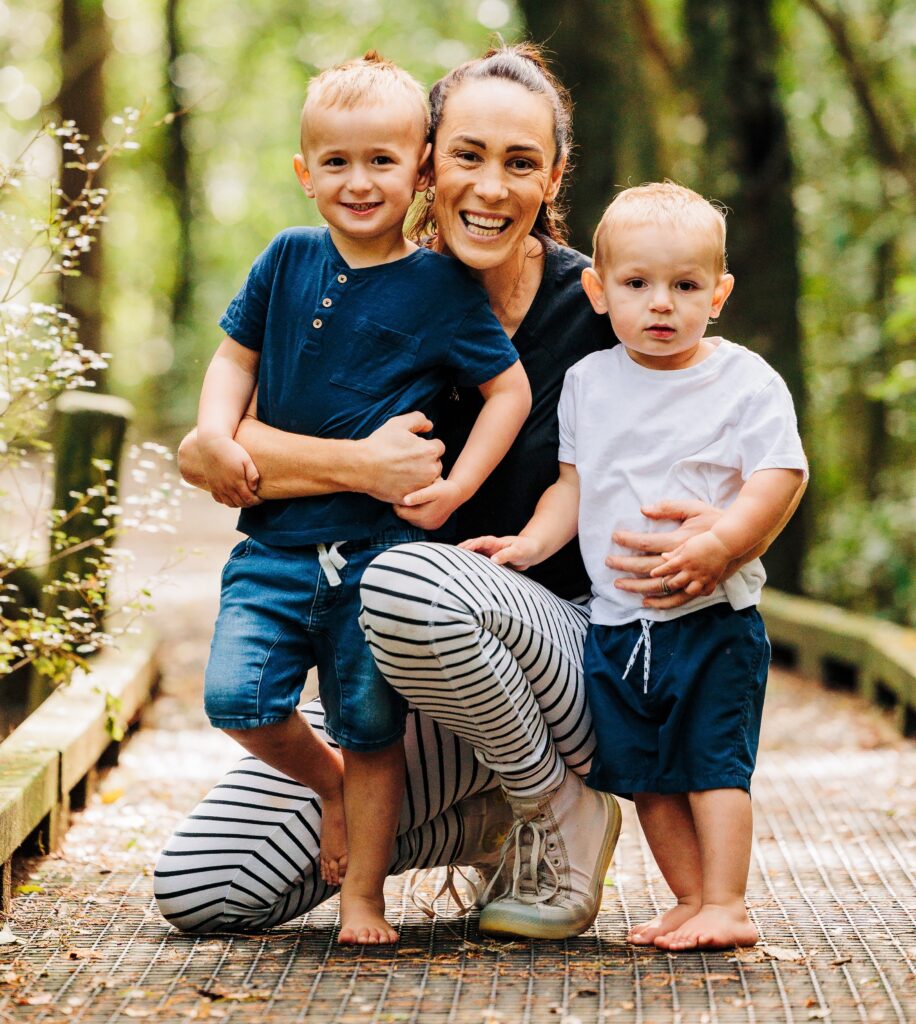 A photo of Lynda Hills, a woman kneeling down, with her arms around two small children, smiling at the camera